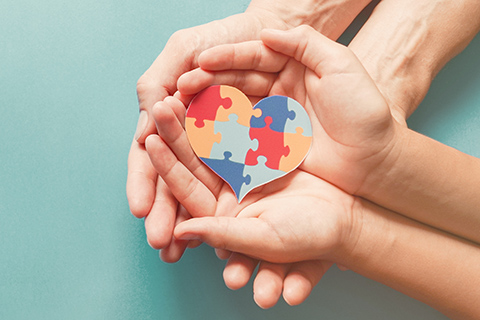 This is a stock photo from Shutterstock. Adult hands are cupped together with a child's hands resting inside of them. The child's hands are cupped. The child is holding a heart put together by multi-colored puzzle pieces.