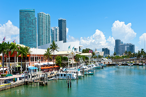 This is a stock photo from Shutterstock. This photo was taken at Bayside Marketplace. This is an outdoor mall that sits on Biscayne Bay in downtown Miami, FL. In the photo, on the left side, there are boats docked in the marina, palm trees, and there are people walking around. There are skyscrapers in the background. On the right side of the photo is the bay.