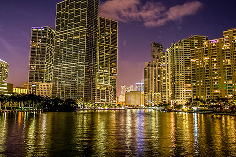 This is a stock photo. This photo was taken at night along the Miami River. This portion of the river is located near the Brickell area of downtown Miami. The view is over the water facing outward towards  the many high-rise buildings. All the buildings are lit up by bright white and yellow lights.