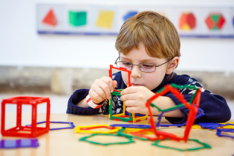 This is a stock photo from Shutterstock. A young boy is sitting at a table playing with puzzle pieces. He is building a colorful puzzle. The boy is wearing a sweater and eyeglasses. 