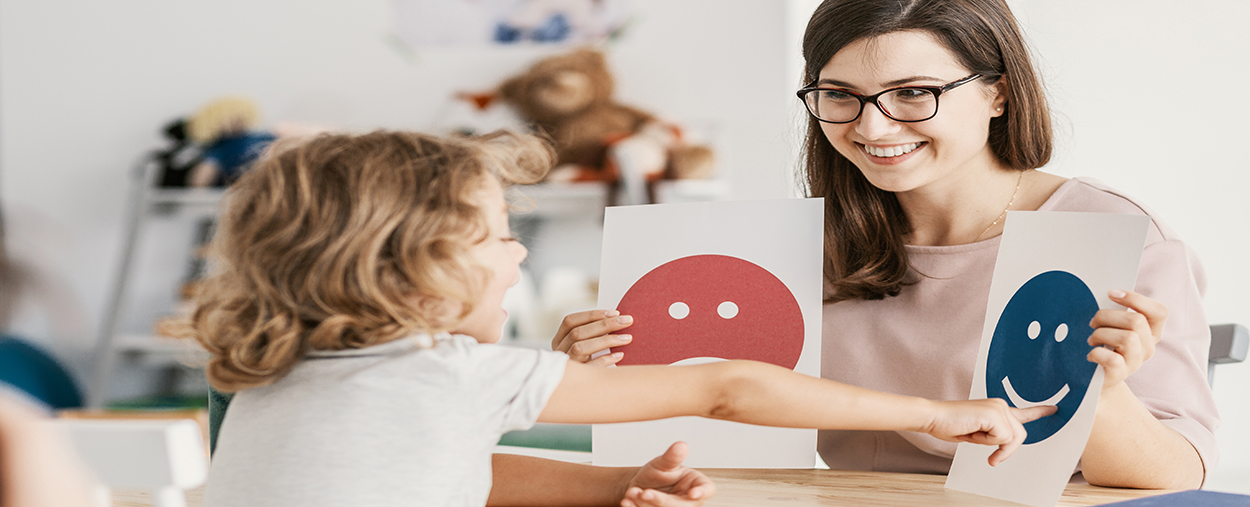 This is a stock photo from Shutterstock. An adult woman is sitting at a table facing a young girl. The woman is holding up two pictures. One picture is a "happy" face, and the other picture is a "sad" face. The young girl is pointing at one of the pictures.
