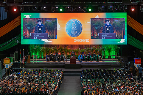 This is an aerial view of the 2022 University of Miami Commencement stage. The university president is speaking at the podium, and the university Dean's are sitting behind him. All of the graduate students are sitting in their chairs facing the stage.