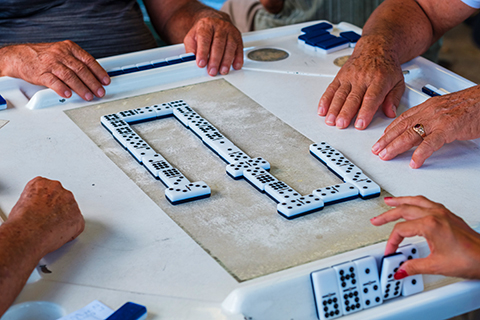 This is a stock photo from Shutterstock. This photo is focused on a game of dominoes. The center of the image shows the domino game with the hands of each player on the edge of the photo. 