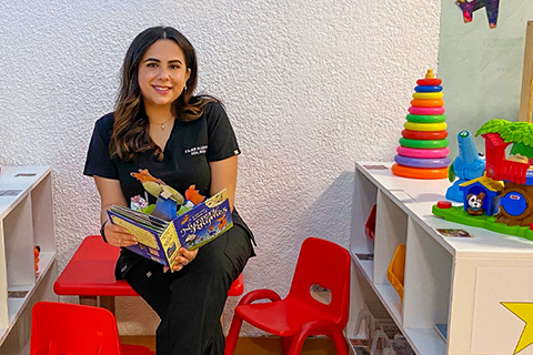 A University of Miami alumnae sits in the clinic she opened for children. Her clinic is modeled after the University of Miami IBIS clinic. She is surrounded by colorful toys and books. She is wearing her clinic uniform.