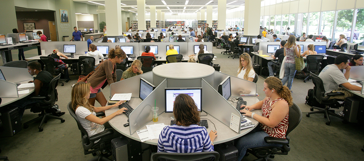 This is a photo inside the University of Miami Richter Library on the Coral Gables campus. The area in the photo is of various individual workstations with computers. This area is busy with many students sitting at the workstations, and some students are walk through the area as well. Some students are at workstations helping their peers at the computer.