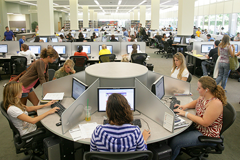 This is a photo inside the University of Miami Richter Library on the Coral Gables campus. The area in the photo is of various individual workstations with computers. This area is busy with many students sitting at the workstations, and some students are walk through the area as well. Some students are at workstations helping their peers at the computer.