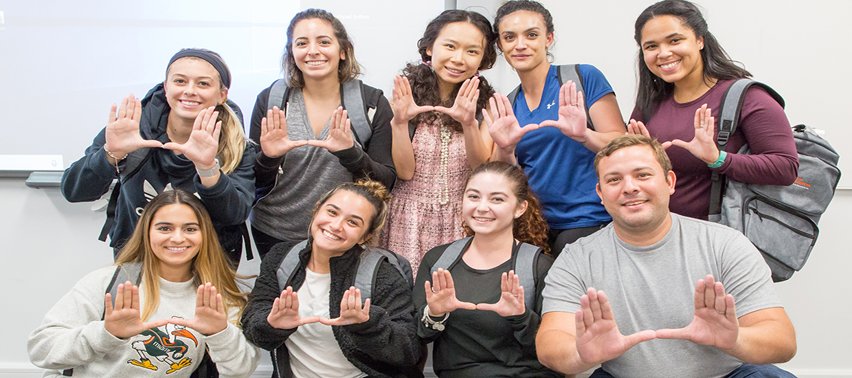 A group of University of Miami students pose for a photo. Each student uses both of their hands to form a "U" shape. The University of Miami "U" is made by placing both hands in front, palms face away from your face and touch the tips of your thumbs together.