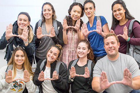 A group of University of Miami students pose for a photo. Each student uses both of their hands to form a "U" shape. The University of Miami "U" is made by placing both hands in front, palms face away from your face and touch the tips of your thumbs together.