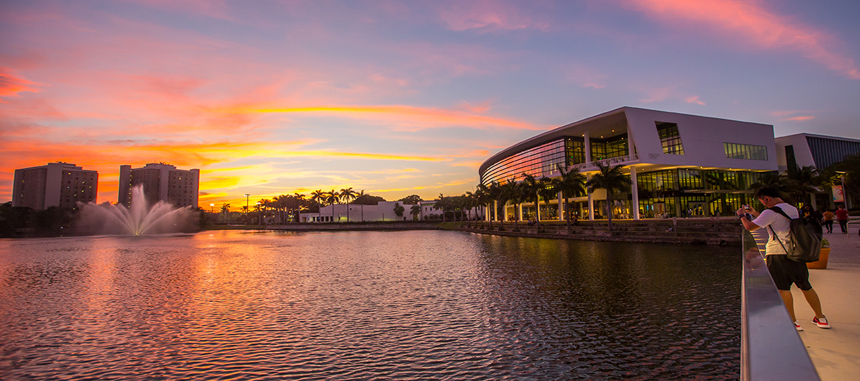 This is a photo of Lake Osceola and the Shalala Student Center at the University of Miami Coral Gables campus. The photo was taken across the lake opposite of the Shalala Student Center. A student is in the foreground using their cellphone to take a picture of the sunset.