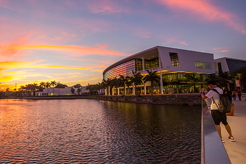 This is a photo of Lake Osceola and the Shalala Student Center at the University of Miami Coral Gables campus. The photo was taken across the lake opposite of the Shalala Student Center. A student is in the foreground using their cellphone to take a picture of the sunset.