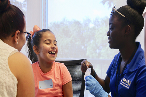 This photo was provided by the University of Miami & Nova Southeastern University Center for Autism and Related Disabilities. A student is speaking with a young patient and her mother.